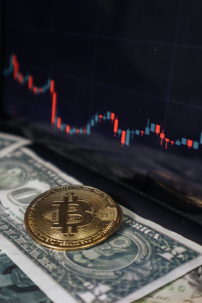 Bitcoin coin resting on a dollar bill with a stock market chart on a screen behind, symbolizing cryptocurrency investment.